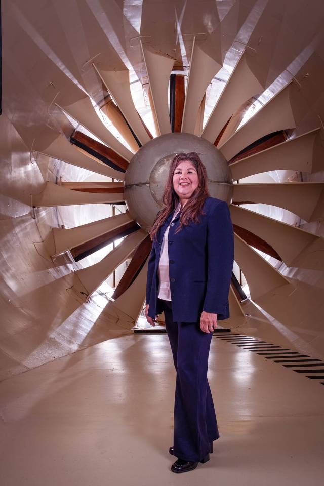 Adabelle Narvaez-Bostwick poses inside NASA Glenn’s Icing Research Tunnel. She is smiling, wearing a blue suit, and standing in front of a large, tan fan.