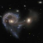 Two merging galaxies. Left one has a large spiral arm curving out from the core, around to below it. Right one has bright core but elsewhere is very faint. A broad curtain of gas connects the two galaxies’ cores and hangs beneath them.