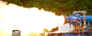 A 24-inch subscale solid rocket motor has a large flame out the left side of the screen during a hot fire test.