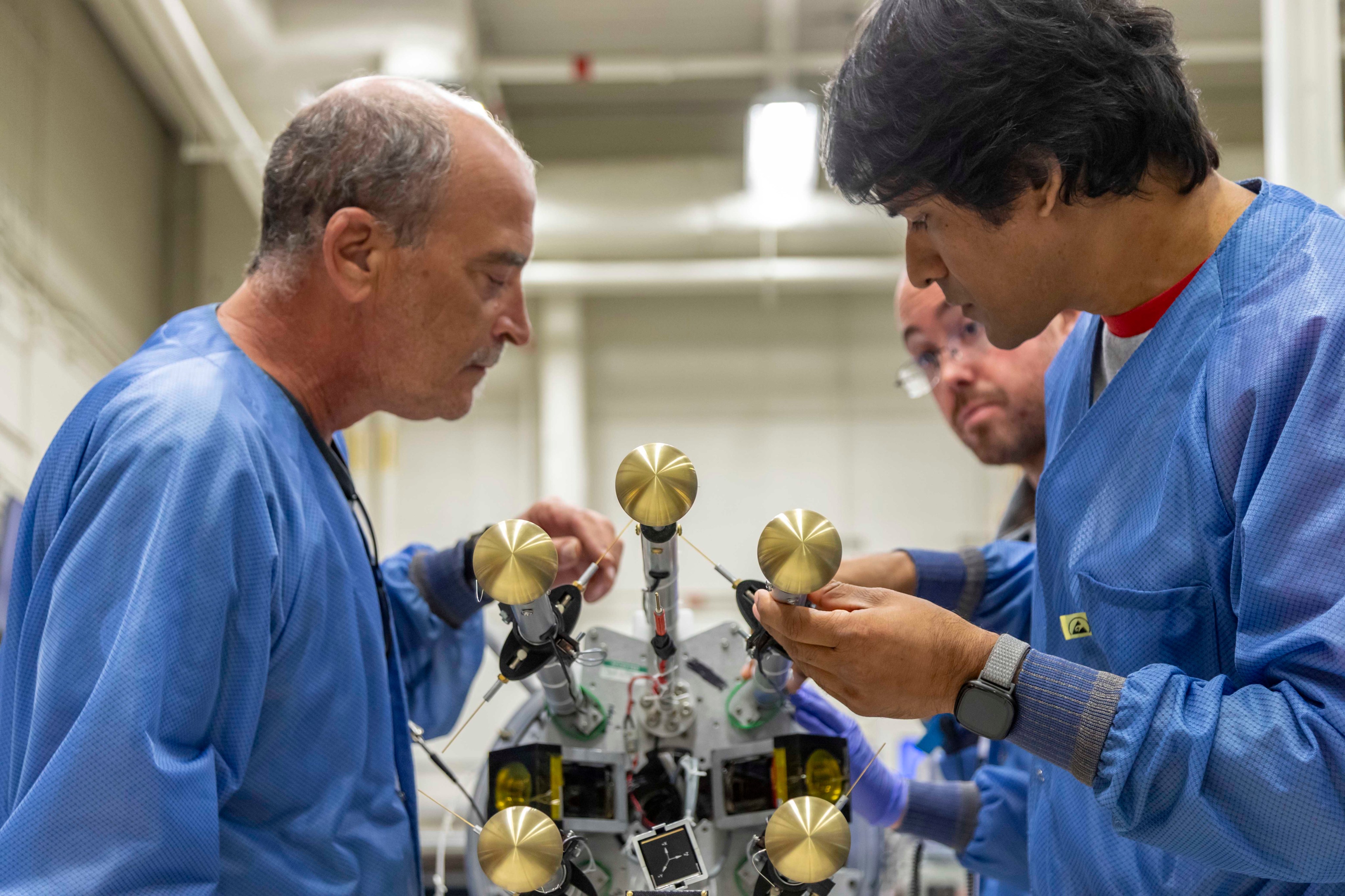 Three men stand over a rocket laid down on a table in front of them. Two in the foreground are adjusting a gold-colored metal rod protruding from the end of the rocket.