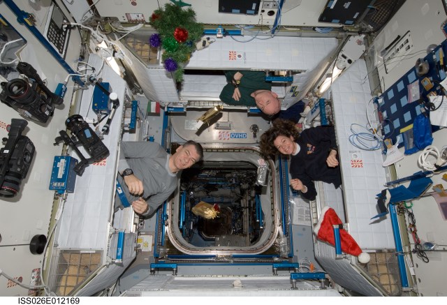 Three of the six crew members aboard the International Space Station peek out of their sleeping quarters to view the station’s decorations and gifts.