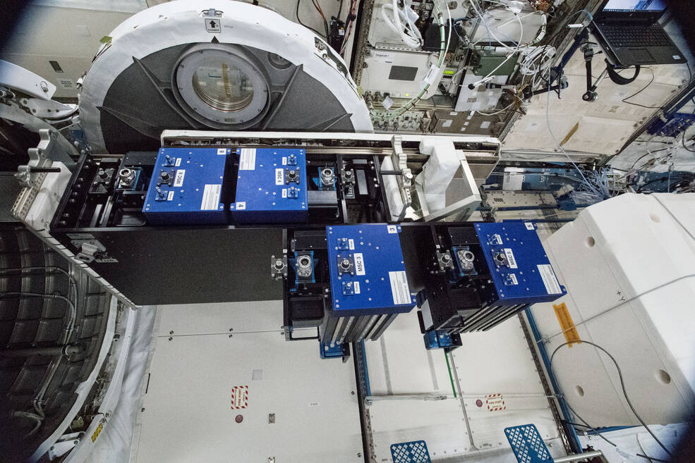 In this image, blue-topped containers are shown from which hang multiple sample cartridges that are staged in the JEM Airlock for installation on MISSE.