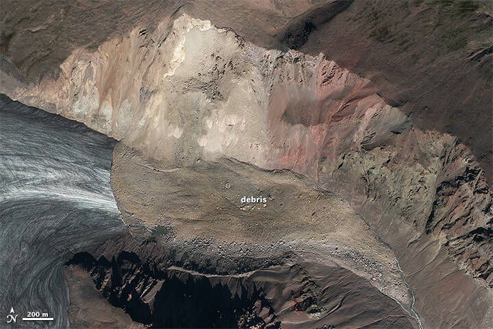 A satellite photo of a landslide in Alaska. The land appears in shades of light and medium brown with gray and pink hues; it cuts away sharply like a cliff face, with light brown debris piled at the foot of the cliff. Gray water is in the lower left.