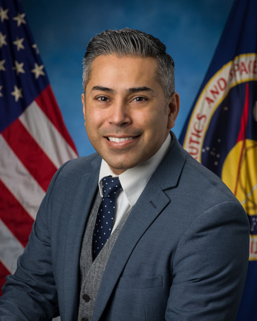 A picture of a man in a suit and tie in front of the United States and NASA flags
