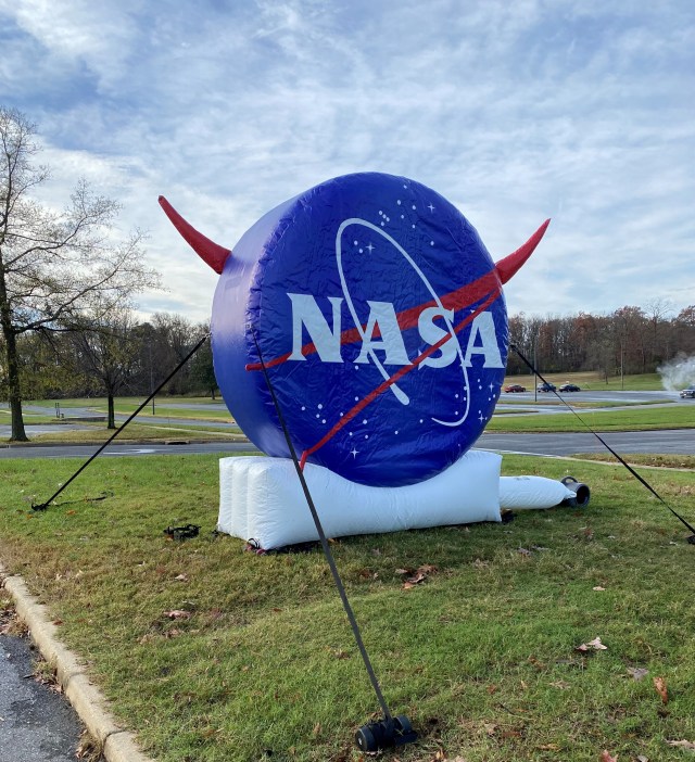 A photo of a large, inflatable NASA logo, which looks like a big blue circle with a red V-shape running diagonally across it, behind big white letters saying NASA and surrounded by stars. The word NASA and the red V are circled by a white oval representing a comet's path. The inflatable has the logo on both sides, so the point of the V on the back side is visible extending out the left side of the circle. The inflatable is staked to the ground outdoors with cables, and it is a slightly cloudy day with green-brown grass and trees in the background.