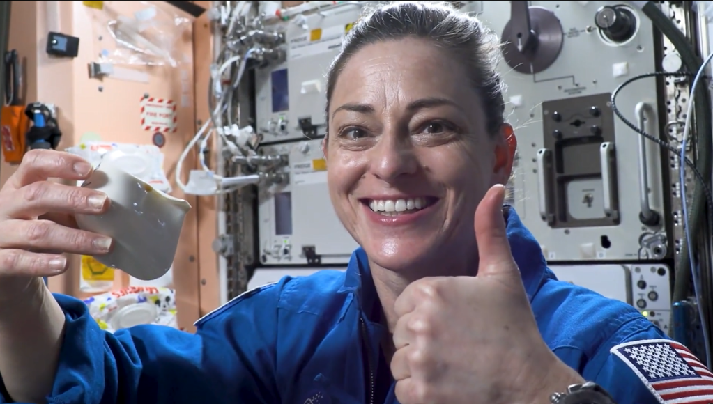 NASA astronaut Nicole Mann holds the Capillary Beverage Cup, or "space cup," aboard the International Space Station while smiling at the camera and giving a thumbs up.