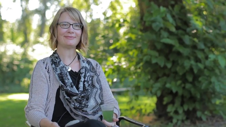 Portrait of Niki Parenteau in a black dress, light brown sweater, and a black/white scarf.