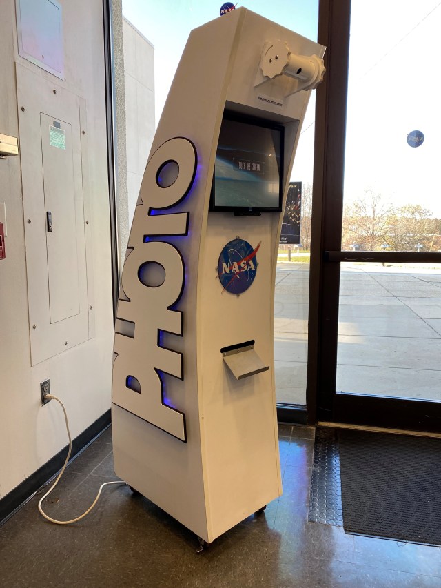 A photo of a photo kiosk designed to take photos of people's faces and insert them into photos of astronauts. The kiosk is taller than a person and shaped like a slightly curved rectangle, with the word "PHOTO" large on the left side and the NASA logo and a screen on the front. A camera is mounted on a bar at the top, and a tray for the printed photos is underneath the NASA logo. The kiosk stands in a brightly lit lobby area, with the outdoors visible through windows behind it.