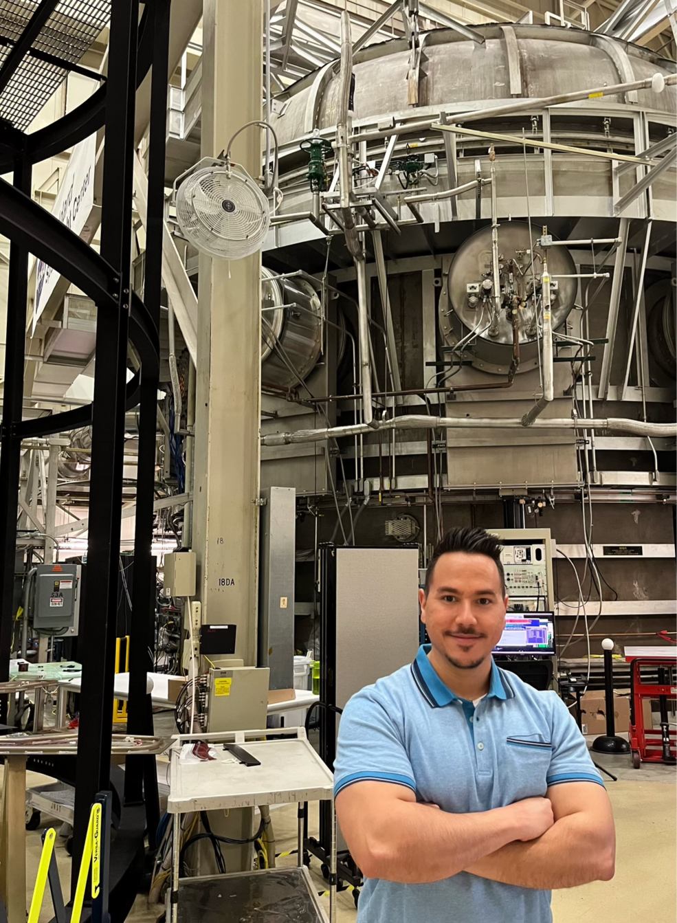 Javier Ocasio-PÃ©rez, a man with dark hair and dark beard and mustache, smiles and stands with arms crossed in front of a test chamber at Goddard, which looks like a large silver cylinder with many silver valves and pipes.