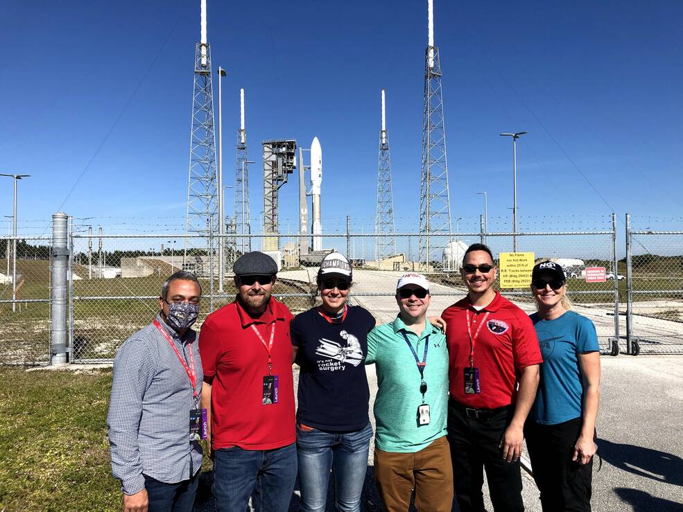 Javier Ocasio-PÃ©rez, a man with dark hair, beard, and mustache, stands with a group of people smiling in front of a rocket on its launch pad on a bright, sunny day. The people are various ages and genders.