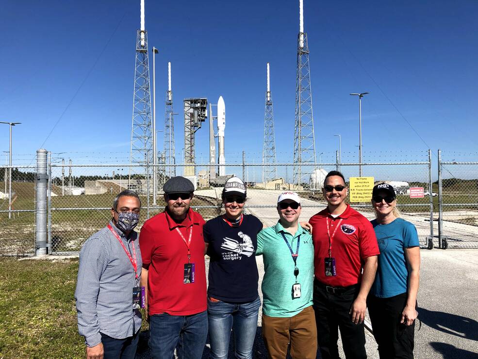 Javier Ocasio-Pérez, a man with dark hair, beard, and mustache, stands with a group of people smiling in front of a rocket on its launch pad on a bright, sunny day. The people are various ages and genders.