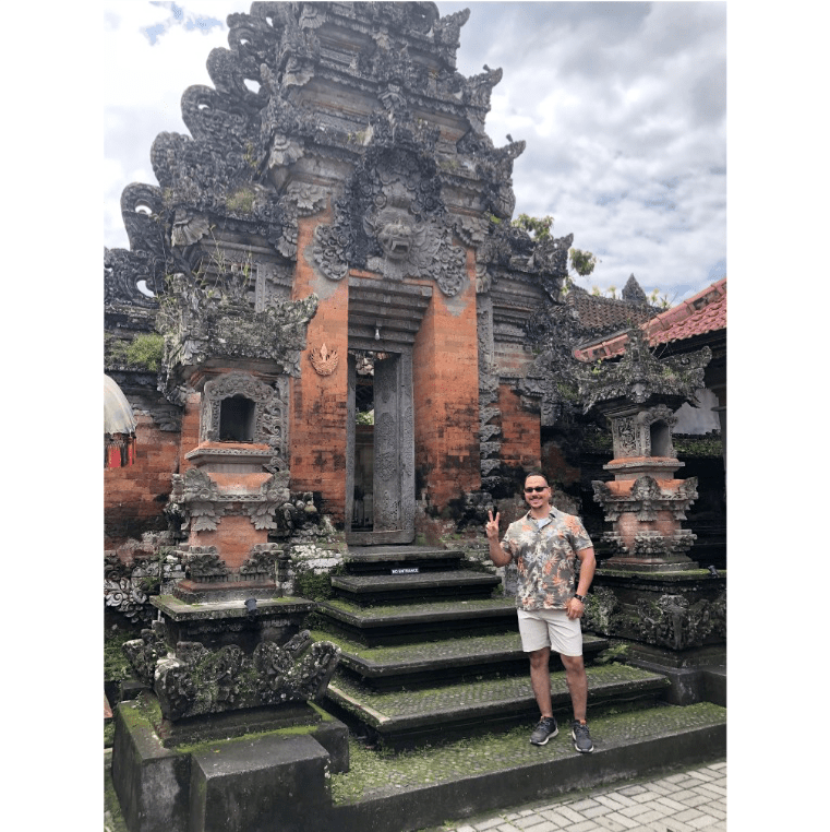 Javier Ocasio-PÃ©rez, a man with dark hair, beard, and mustache, smiles and poses with one hand up in a peace sign next to an elaborate stone temple. The temple is shades of gray and brown with moss-covered carvings.