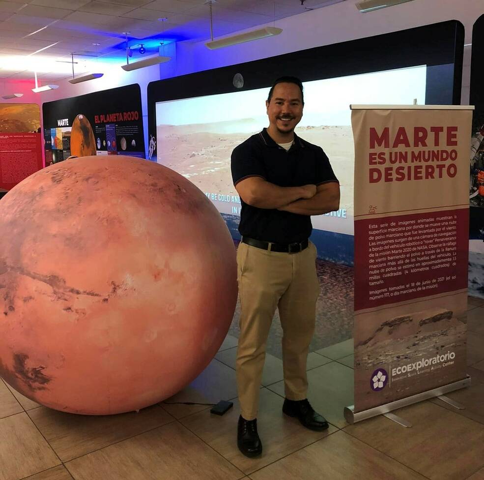 Javier Ocasio-Perez, a man with short dark hair and a dark mustache and beard, smiles and stands with arms crossed next to a large model of Mars. Spanish signs and displays about Mars are around and behind him.