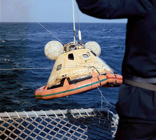 A brownish-yellow space capsule is hoisted out of the ocean. Orange, ball-shaped flotation bags are attached to the top of the capsule, and another orange flotation collar is attached to its bottom.