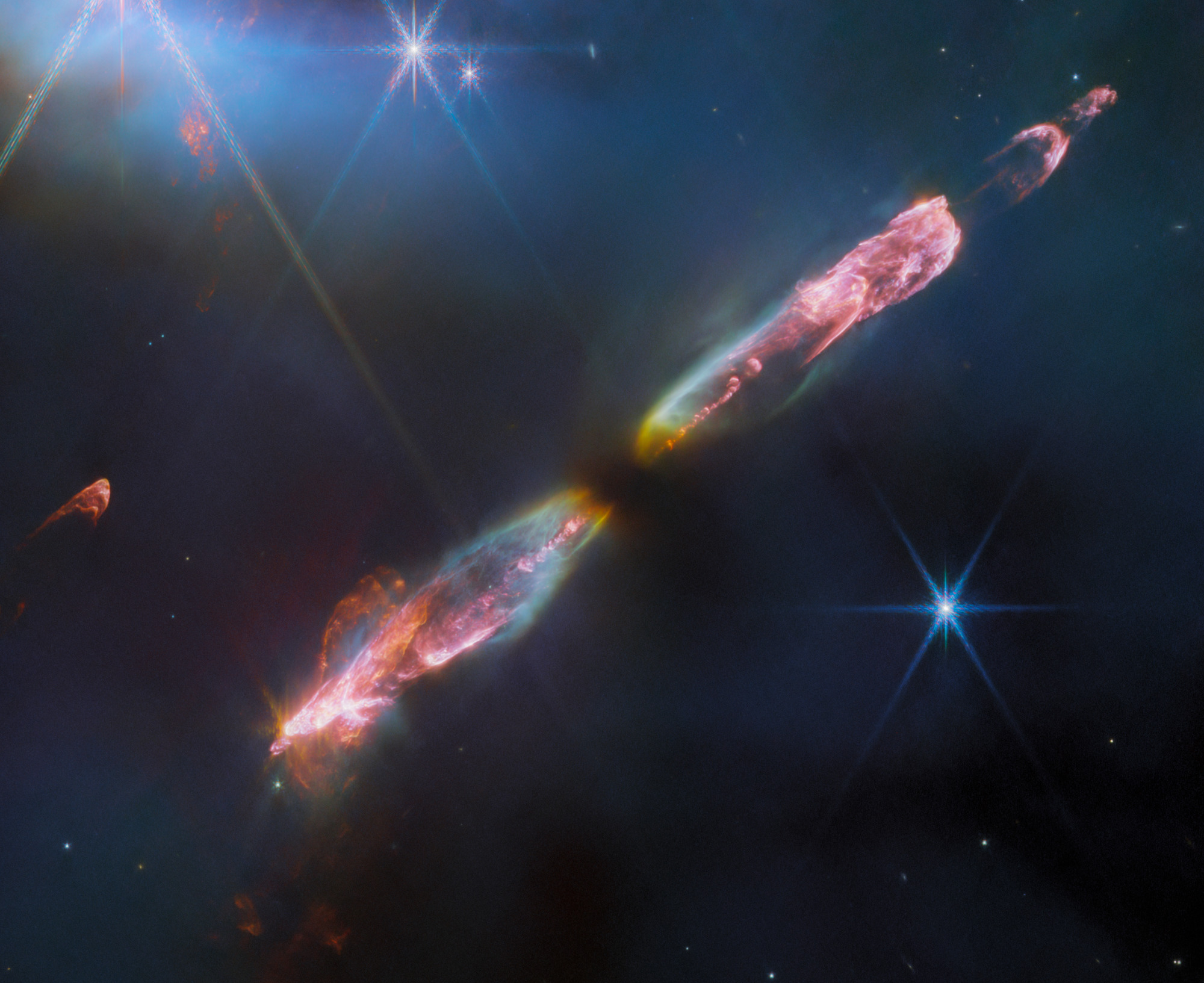 At the center is a thin horizontal pinkish cloud known as Herbig-Haro 211 that is uneven with rounded ends, and tilted from bottom left to top right. It takes up about two-thirds of the length of this angle, but is thinner and longer at the opposite angle. At its center is a dark spot. On either side of the dark spot, there are orangish yellow wisps that extend to light blue wisps. Within the center of those clouds, a pink fluffy streak runs through each lobe. At the ends of each lobe, pink becomes the dominant color. The lobe to the left is fatter. The right lobe is thinner, and ends in a smaller pink semi-circle. Just off the edge of this lobe is a slightly smaller pink semicircle, then a pink sponge-like blog. The background contains several bright stars, each with eight diffraction spikes extending out from the central bright point.