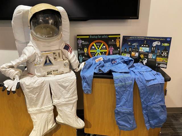 A photo of two astronaut suits draped on a table, with the legs hanging over the edge. On the left is a white suit with an attached helmet and pack on the back. On the right is a blue flight suit. Both suits have many patches and insignia on them. Display cards with graphics and information are propped behind the blue suit, and a TV is mounted on the wall behind them.