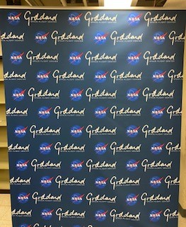 A photo of a banner designed to serve as a background for photo shoots. The banner looks like a curtain hanging from a metal rod, printed with the NASA logo and white text saying Goddard Space Flight Center in a repeating pattern. The banner is slightly glossy and reflects some light near the top.