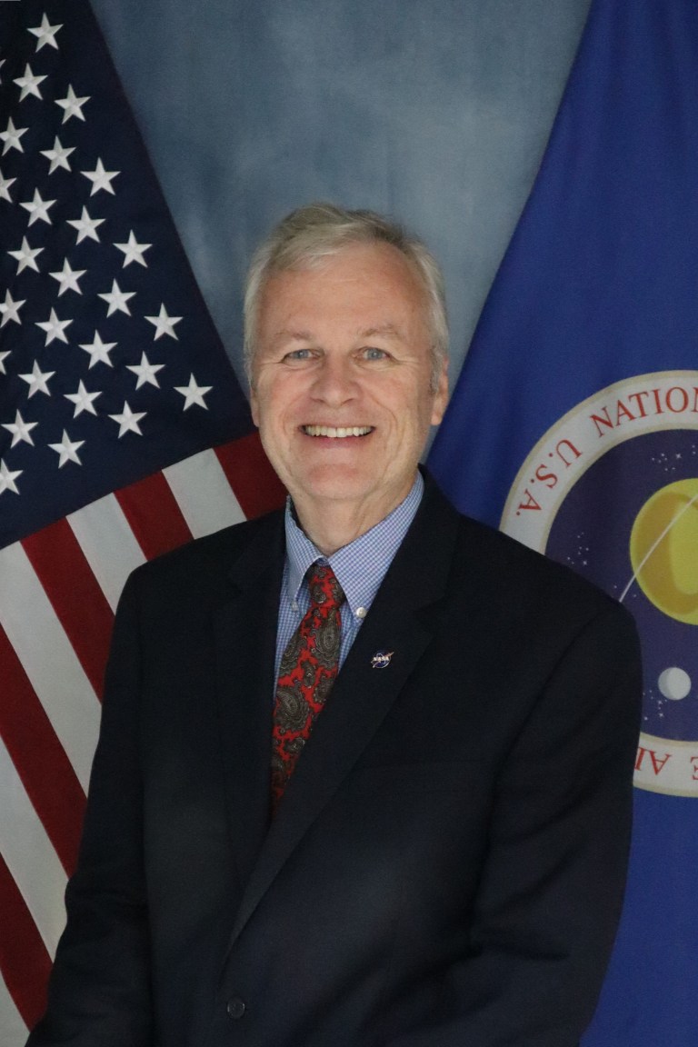 Image shows a NASA employee dress in a suit posing in front of the American flag and NASA flag