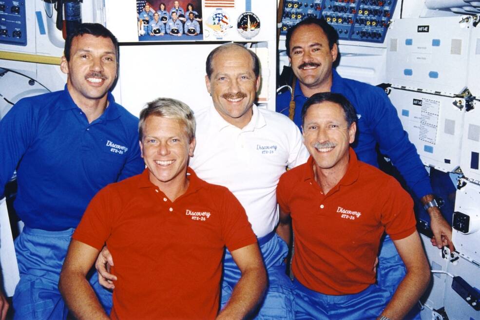 Dressed in red, white, and blue polo shirts, the STS-26 crew poses during the inflight press conference