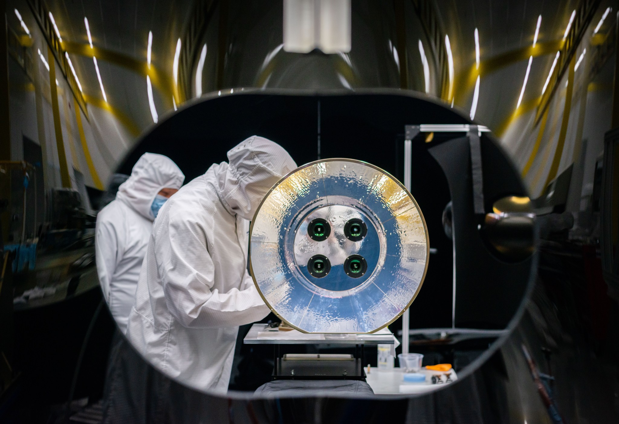 Two scientists wearing white lab coats, hoods, and masks work behind a large reflective disc with four camera lenses in its center, part of the assembly for the AWE.