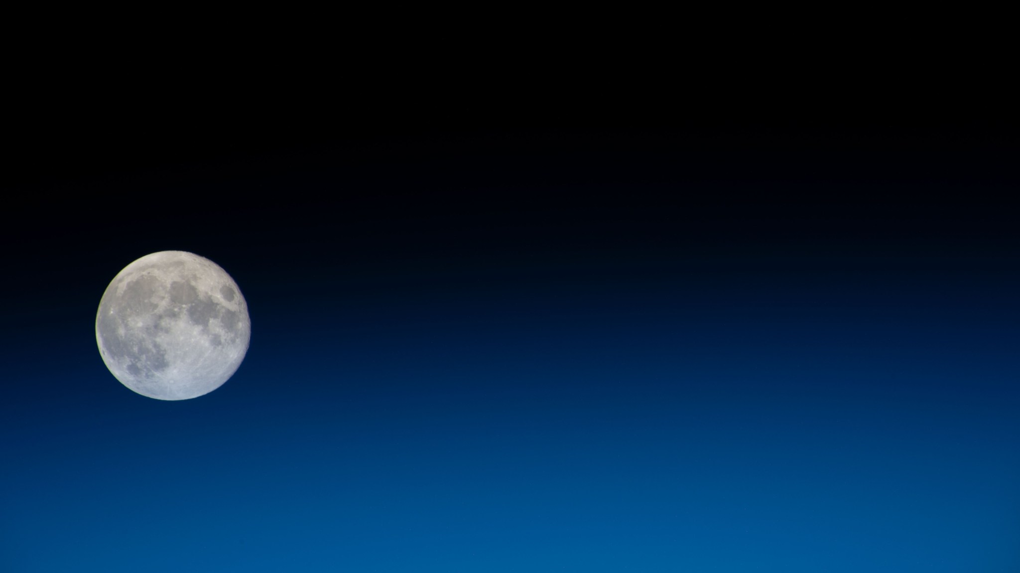 iss070e001516 (Sept. 30, 2023) -- A full Moon is pictured from the International Space Station. The Moon lingers to the left of the image, with a horizon of Earth's blue glow splitting the image nearly in half, blending into the black of space.