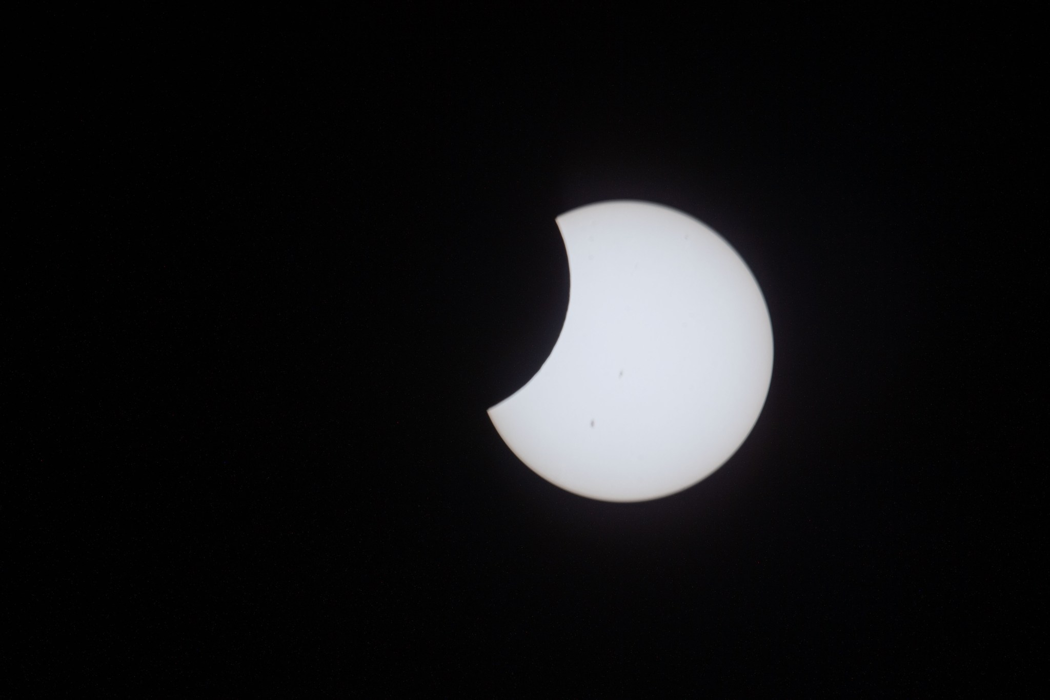 The Sun appears as a white disc in the darkness of space. The Moon begins to pass in front of it on the left, making it look like there's a circular portion of the Sun missing.