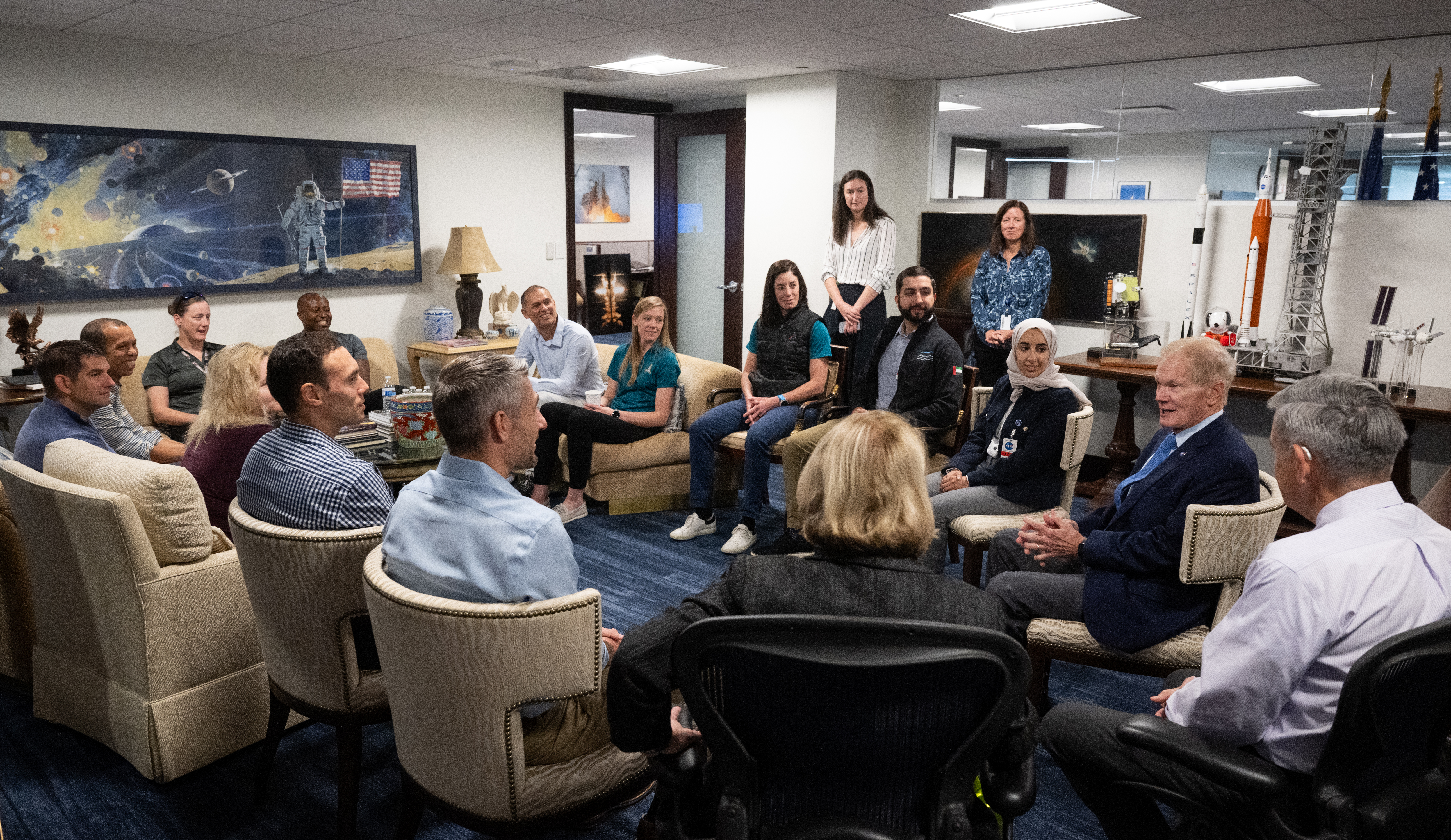 A group of 17 people sit and stand in a circle in a meeting area in an office. There are rocket models and space-themed imagery decorating the space. Everyone looks toward NASA administrator Bill Nelson, second from right, as he speaks.