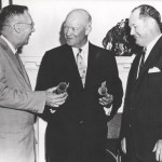 From left to right, Dr. Hugh L. Dryden, President Dwight D. Eisenhower, and Dr. T. Keith Glennan smile as they have a discussion. Dryden and Eisenhower hold cylindrical objects in their hands; Glennan looks down at their hands. They are all wearing suits.