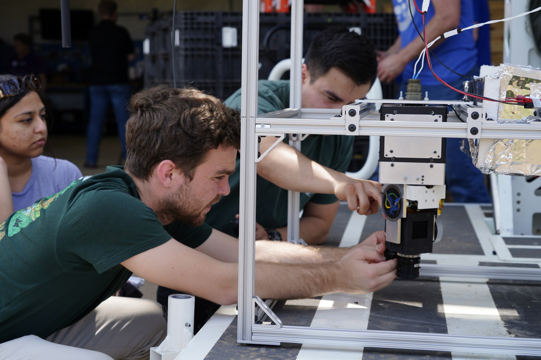 Two people reach into frame-like structure that contains box-like mechanical equipment; one person in background watches integration.