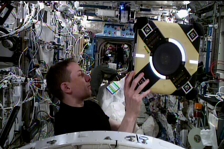The interior of the International Space Station, where an astronaut holds a a cube-shaped robot.