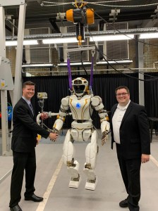 As part of a reimbursable Space Act Agreement with Woodside Energy in Perth, Western Australia, NASA plans to use a Valkyrie robot to develop remote mobile dexterous manipulation capabilities to accommodate remote caretaking of uncrewed and offshore energy facilities. Pictured here from left, Oliver Bentley (US Department of Commerce), Valkyrie, and Evan Laske (NASA). Credits: NASA