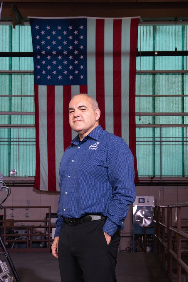 Carlos Garcia-Galan poses in front of the American flag in the Electric Propulsion and Power Laboratory at NASA’s Glenn Research Center. He has a serious expression is wearing a blue long-sleeve shirt with the Artemis program logo and black pants.