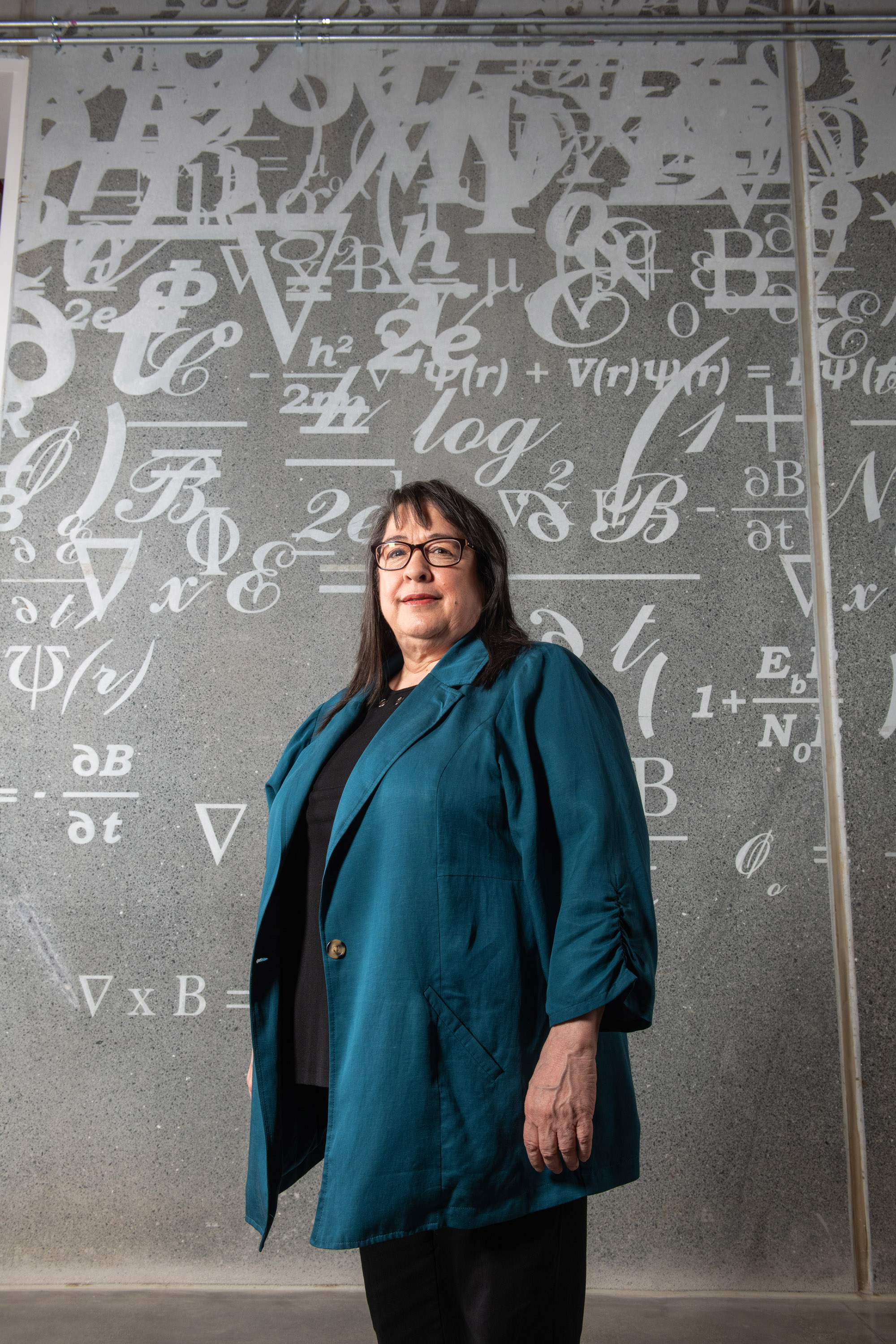 Maricela Lizcano stands inside NASA Glenn Research Center’s Aerospace Communications Facility. She is wearing a teal jacket and behind her there are mathematical equations on the wall.