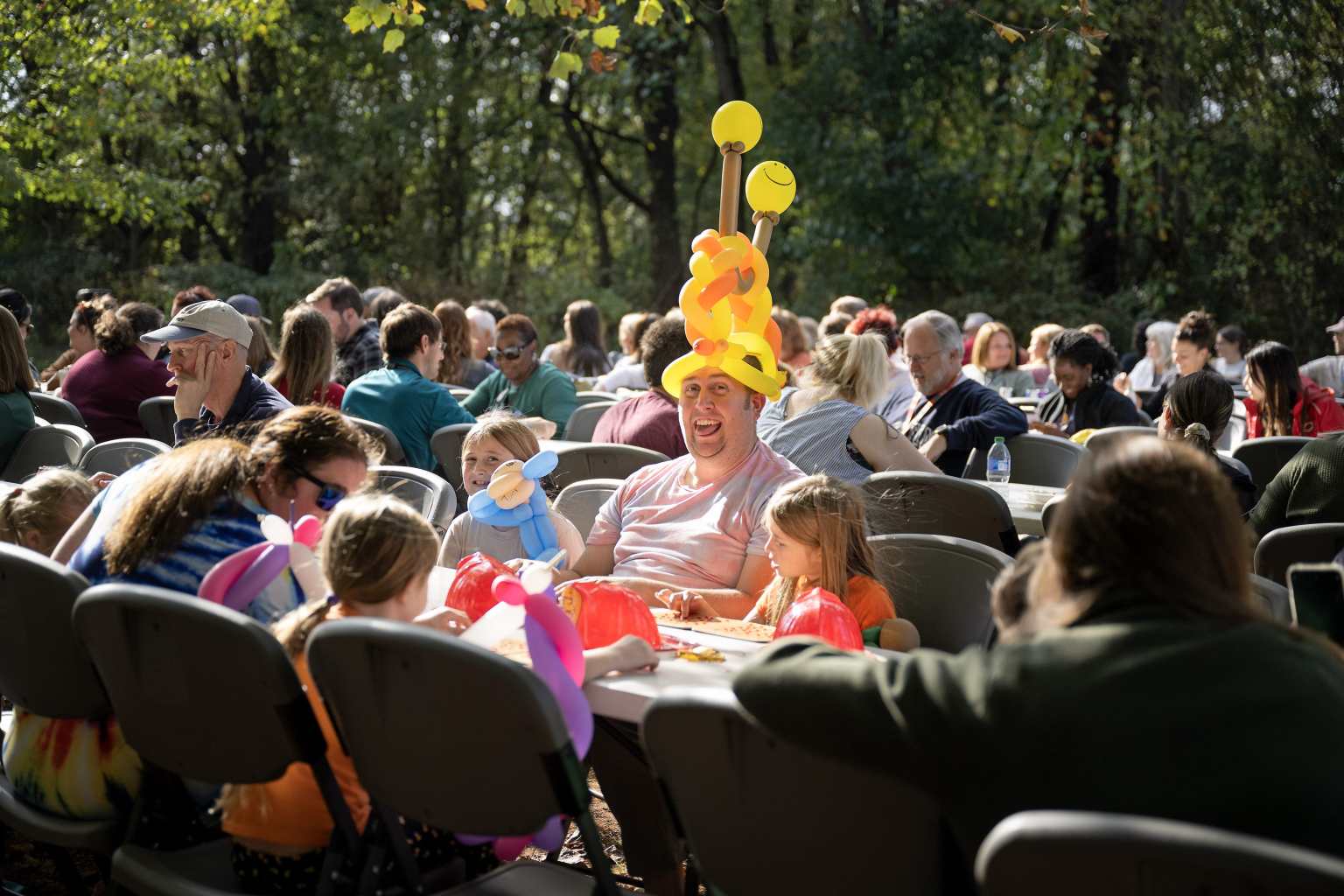 A man with an open-mouthed smile wears a balloon hat made at the Fall Family Fest alongside children who are also enjoying their balloon toys during a Bingo round.