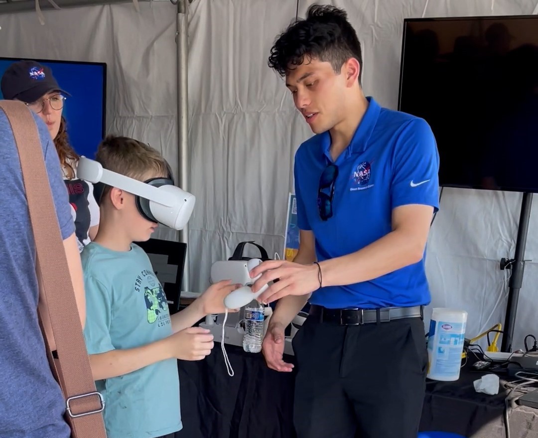 Mark Frances shows a young boy how to use his goggles and controls to experience interactive technology. They are inside a tent.