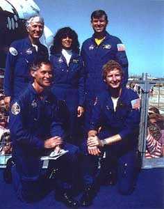 Five of the six STS-41D crew members, Richard M. “Mike” Mullane, kneeling left, Steven A. Hawley, Henry W. “Hank” Hartsfield, standing left, Judith A. Resnik, and Michael L. Coats, pose with Discovery as a backdrop