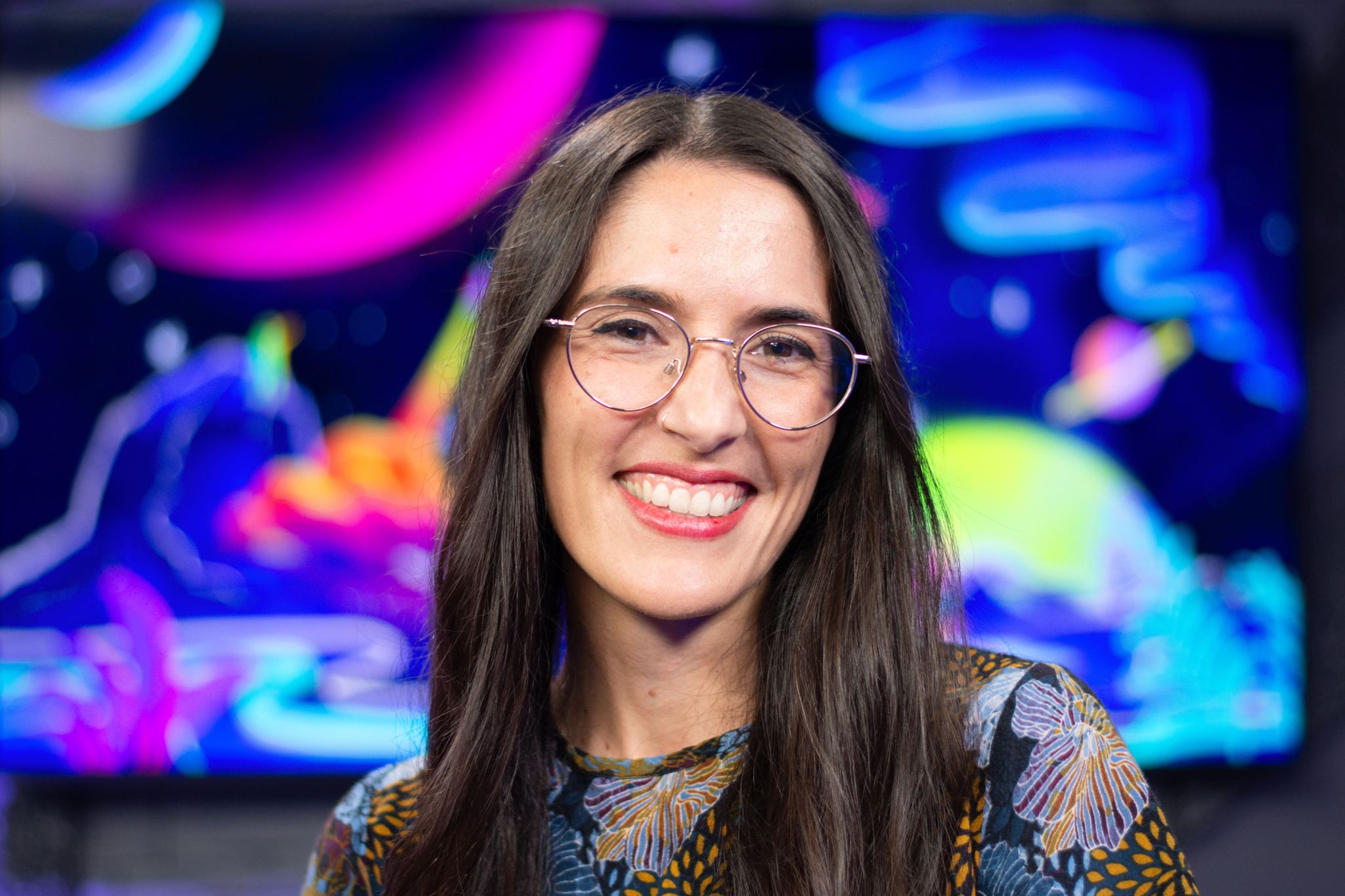 Noelia González smiles widely in the center of the frame. She is wearing large round glasses and a multicolored top of blues and oranges with a flower pattern, and her dark brown hair frames her face. Behind her is the brightly colored artwork for "Universo Curioso de la NASA," NASA’s first-ever Spanish podcast.