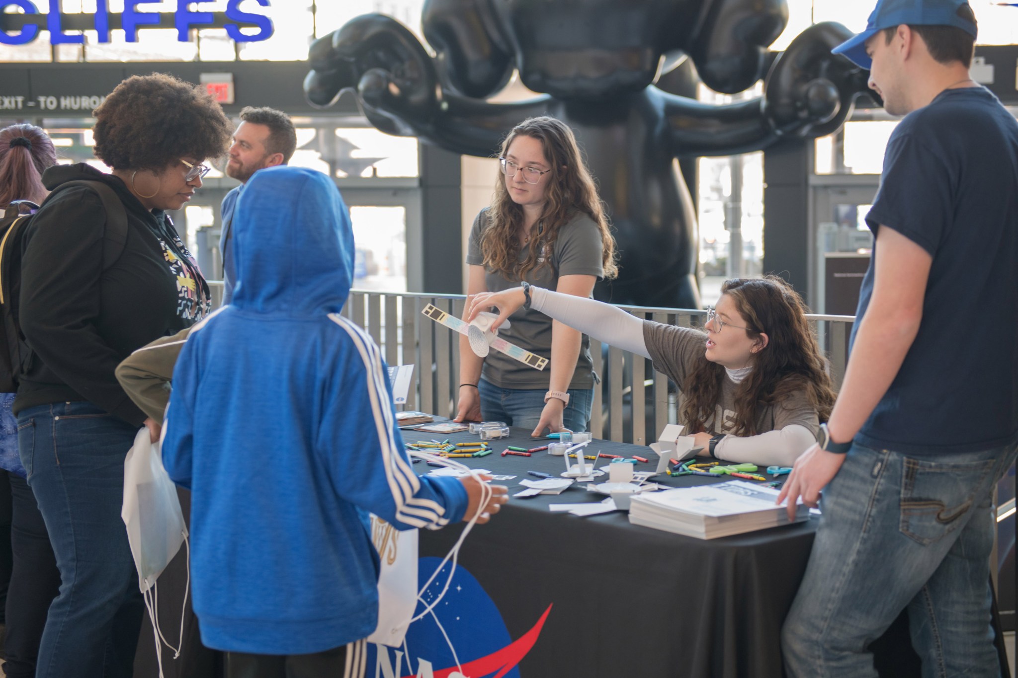 NASA Glenn researchers build paper satellites with students during a STEM event. Two Glenn employees are behind a table, one sitting and one standing. The one sitting holds a paper satellite, and the table is scattered with crayons, paper, and brochures. Three people on the other side of the table watch the demonstration, including a child in a blue hoodie who faces away from the camera.