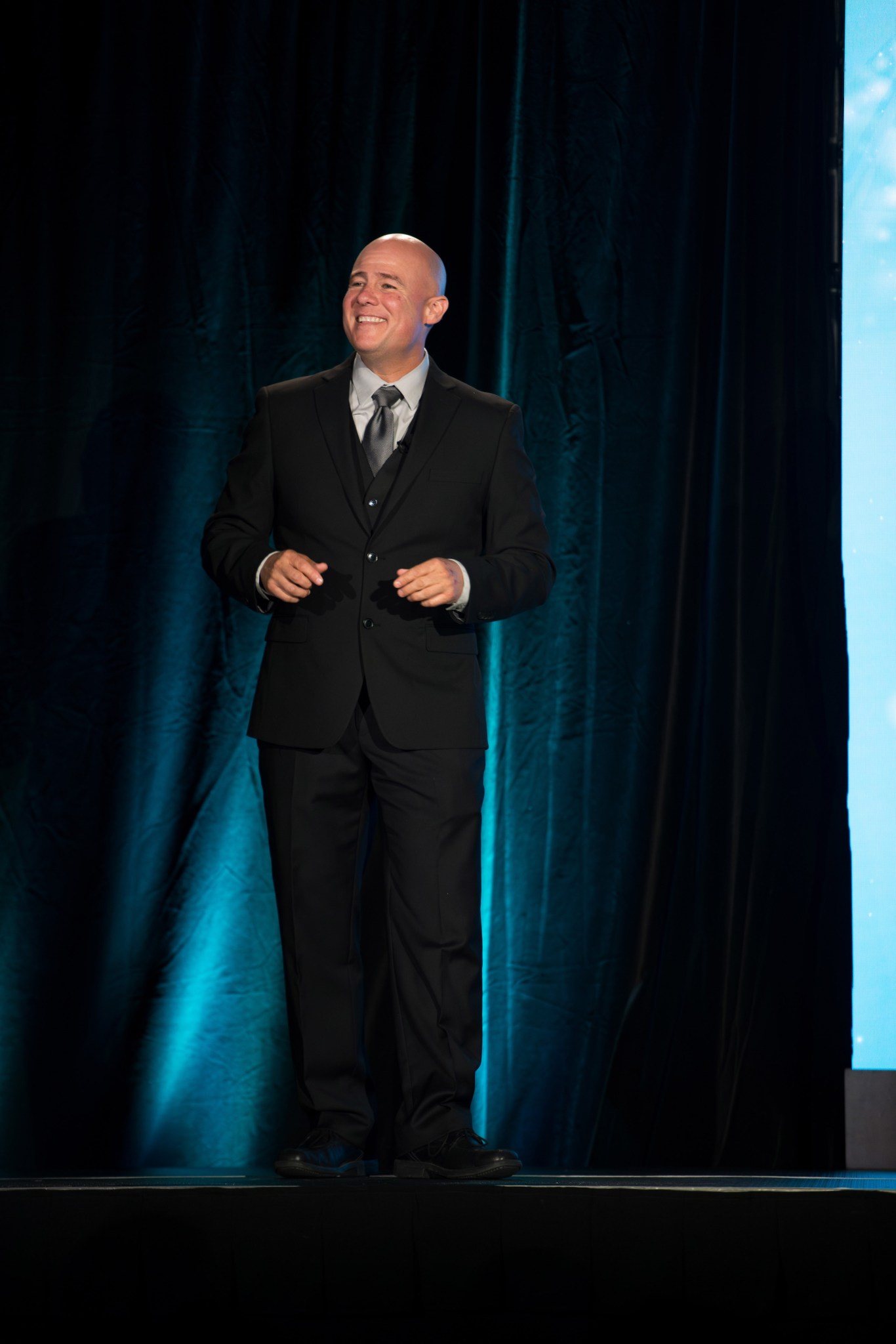 Carlos Flores, in a suit, smiles as he addresses an audience from a stage.