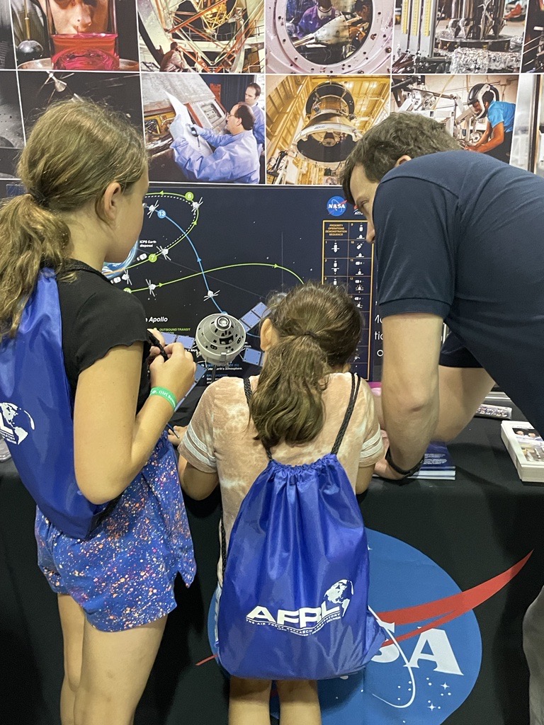 Michael Belair, with two small girls at either side, explains information at a table with colorful images of space. Their backs are facing the camera.
