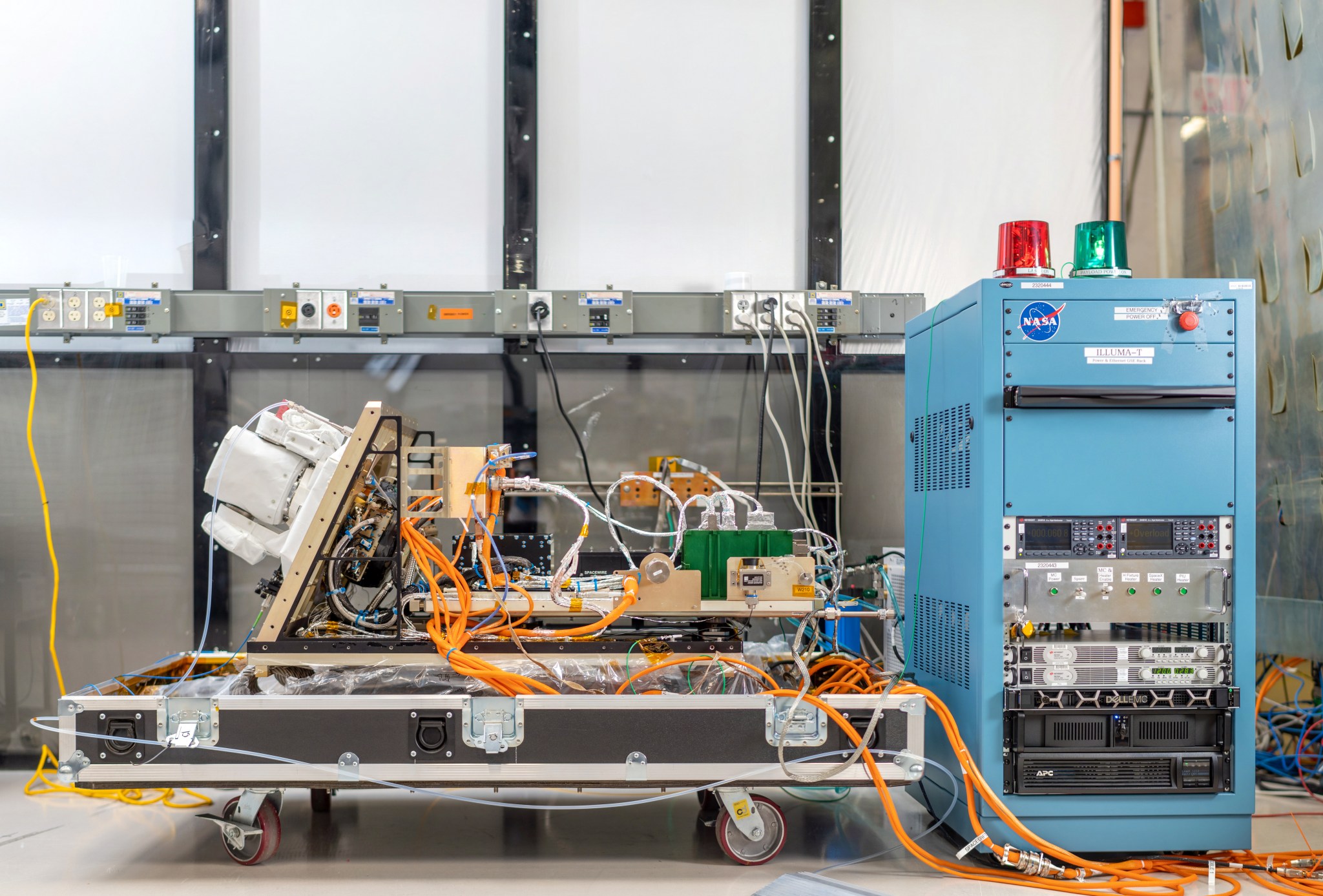 Hardware including tangles of large orange cords and smaller white cords connecting various boxes and boards sits on a large, wheeled cart. To the right is a refrigerator-sized blue computer box topped with red and green flashing lights. A power strip runs along the wall behind.