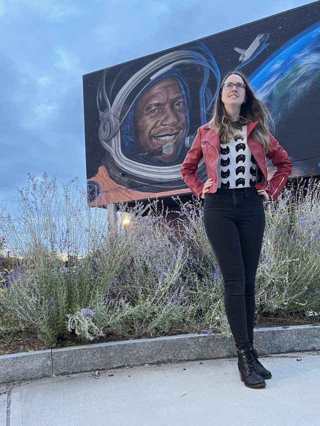 Sarah Tipler poses in front of a mural of NASA astronaut Michael Anderson in Plattsburgh, New York. She is wearing a red jacket and a white shirt with black cats on it and is looking toward the sky. The colorful painted mural features Anderson wearing an orange spacesuit, the shuttle Columbia, and a view of Earth. The sky is blue and grass with purple flowers can be seen in front of the mural and behind Tipler.