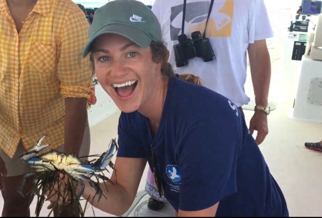 Shaigh Sisk, a woman with curly brown hair pulled back in a braid, smiles joyfully at the camera while holding a large blue and yellow crab in her right hand. She wears a green hat and navy tee, and is kneeling and holding the crab. Other people in yellow and white shirts stand behind her.