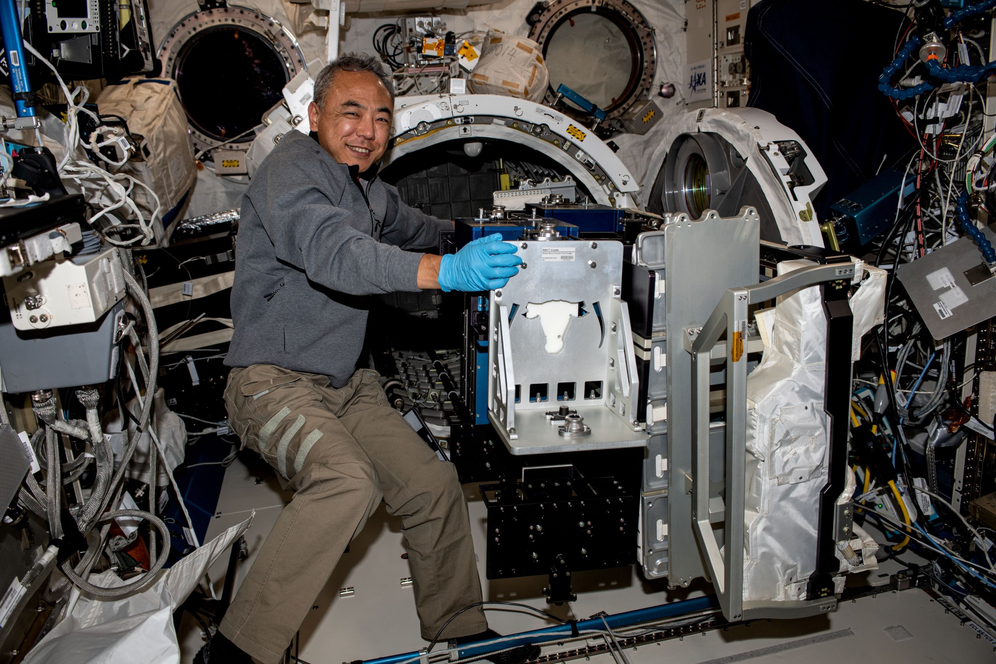 Furukawa, wearing a gray shirt and khaki pants, smiles at the camera as he pulls hardware through the open cylindrical door of an airlock. The suitcase-sized hardware has a silver front, with blue boxes behind it.