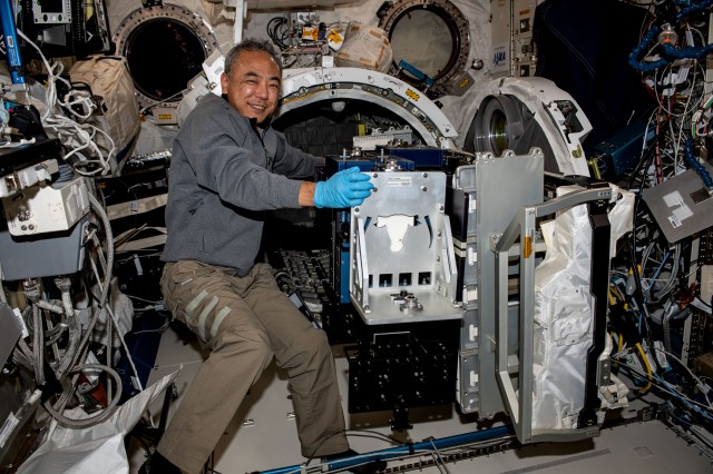 Furukawa, wearing a gray shirt and khaki pants, smiles at the camera as he pulls hardware through the open cylindrical door of an airlock. The suitcase-sized hardware has a silver front, with blue boxes behind it.