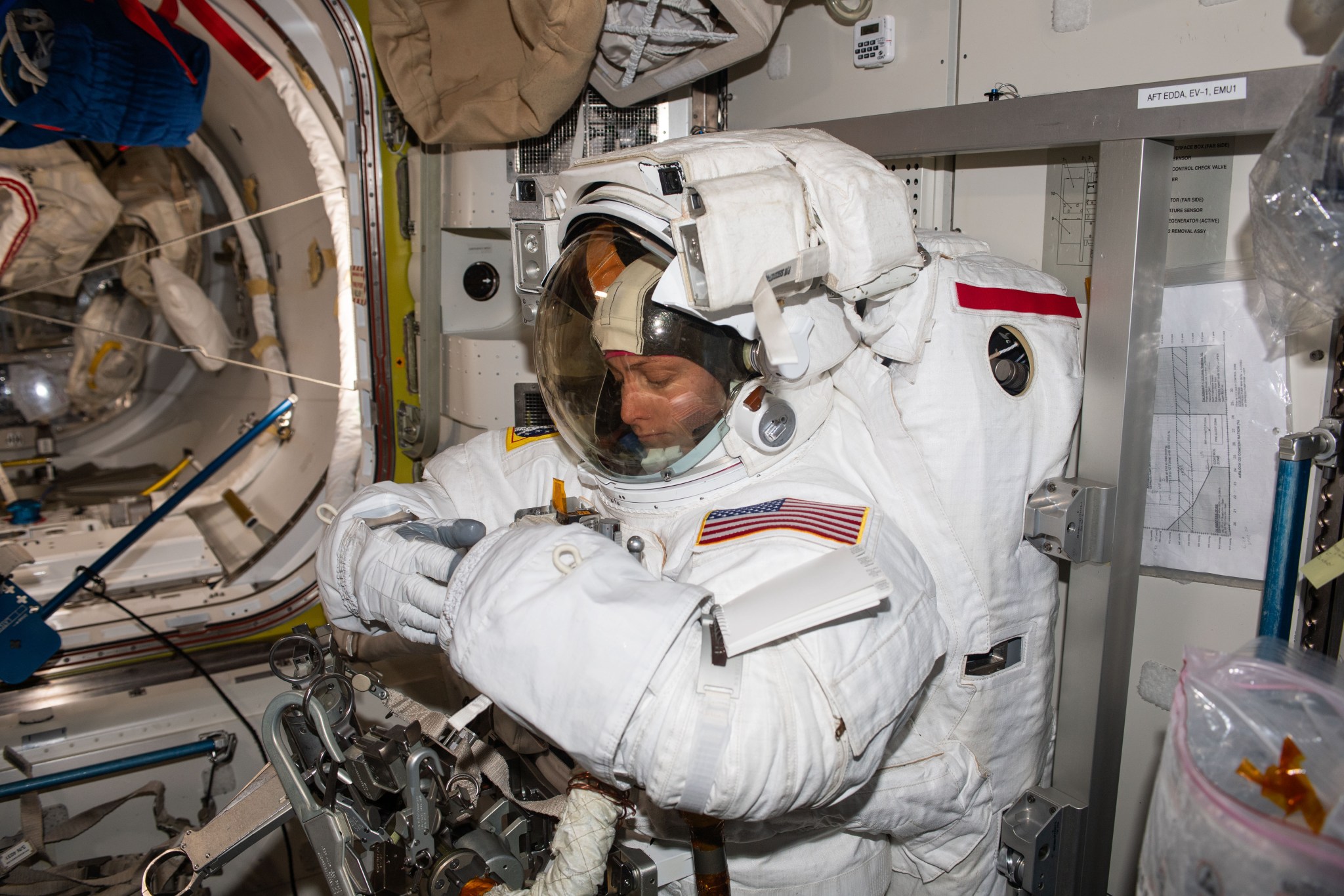 O’Hara is inside a white spacesuit with an American flag on the shoulder. Her face is visible through the clear front of the helmet, and she is using her gloved right hand to tuck the suit into the glove of her left hand. The suit is attached to a metal frame on the wall.