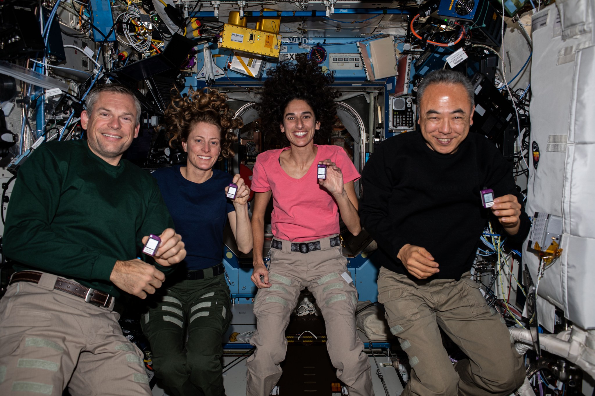 Mogensen is wearing a long-sleeved green shirt and khaki pants, O'Hara a short-sleeved blue shirt and green pants, Moghbeli a short-sleeved pink shirt and khaki pants, and Furukawa a long-sleeved black shirt and khaki pants. All four are smiling at the camera and holding small pager-sized devices that are white on the front and purple around the edges.