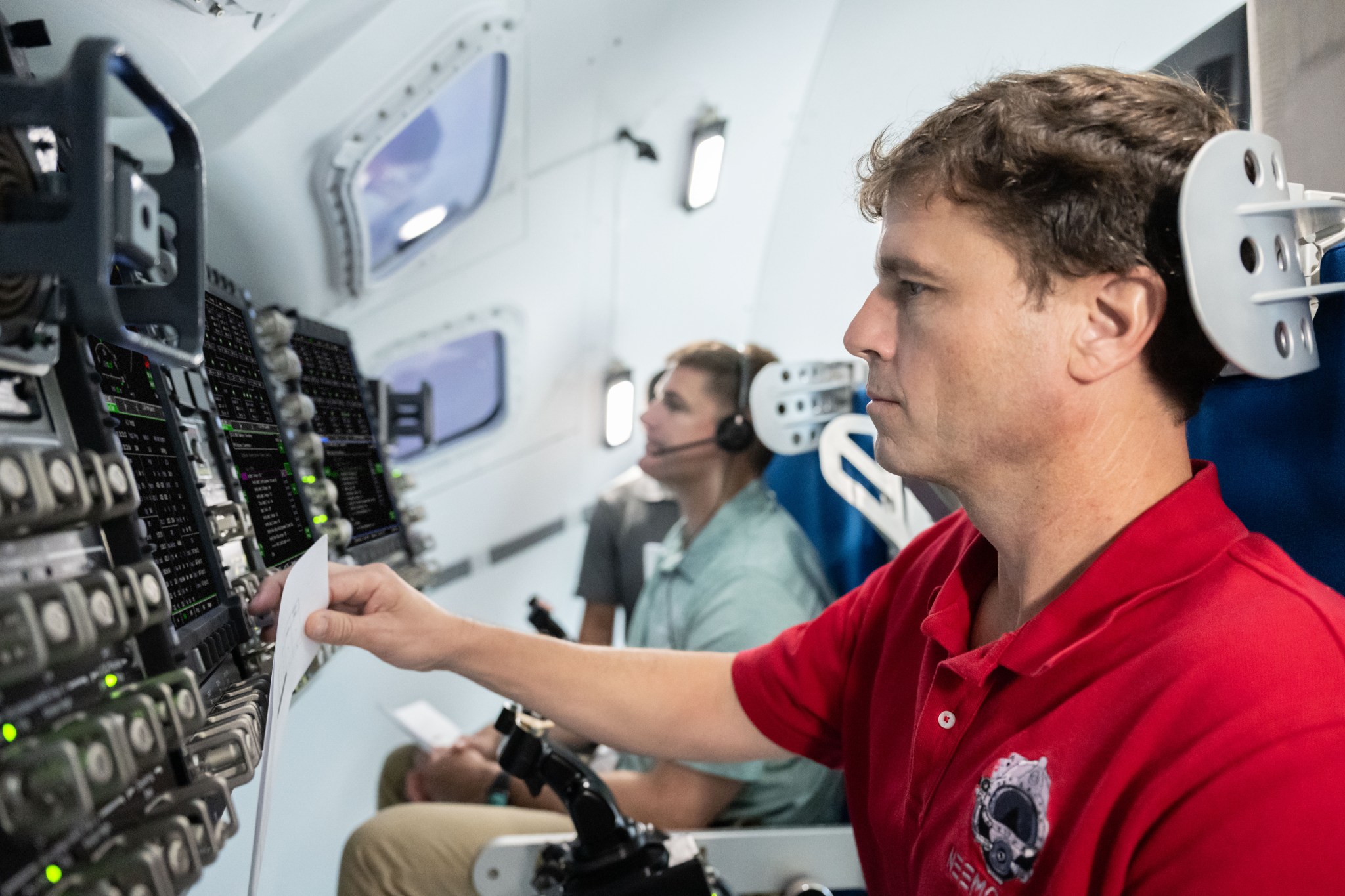 Astronaut Reid Wiseman is in a red shirt in the foreground with his right hand touching Orion's controls while Jeremy Hansen sits in the background.