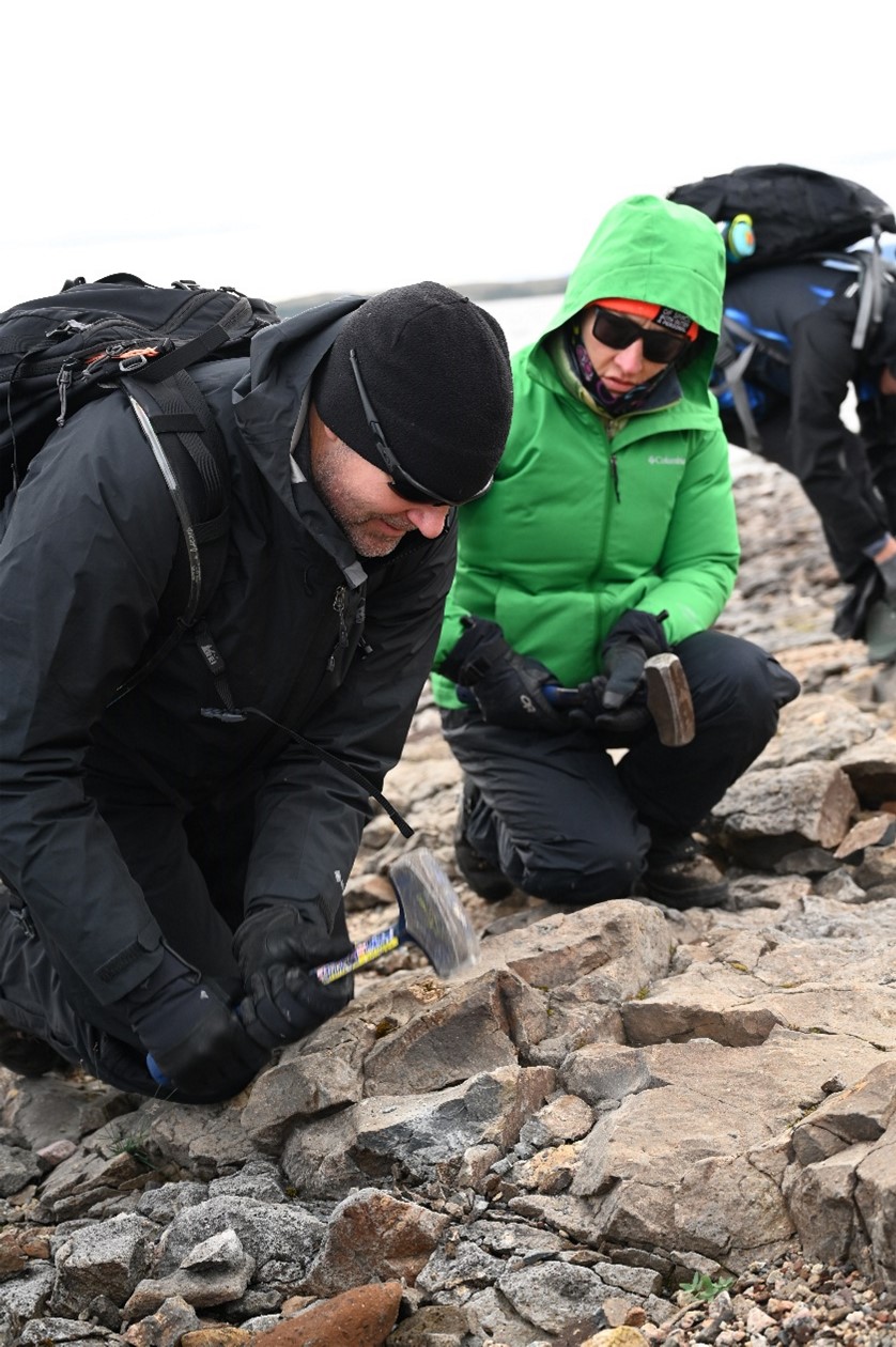 Two astronauts, one in a black jacket and black hat, and the other in a lime jacket with its hood up, kneel to examine the rocky landscape.