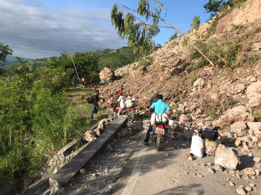 A man on a motorcycle is blocked by a landslide that has fallen across the road,  covering it in large boulders, rocks and debris. A few other men working their way around the blockage. The sky is blue and slightly cloudy, and they are in a forested area. 